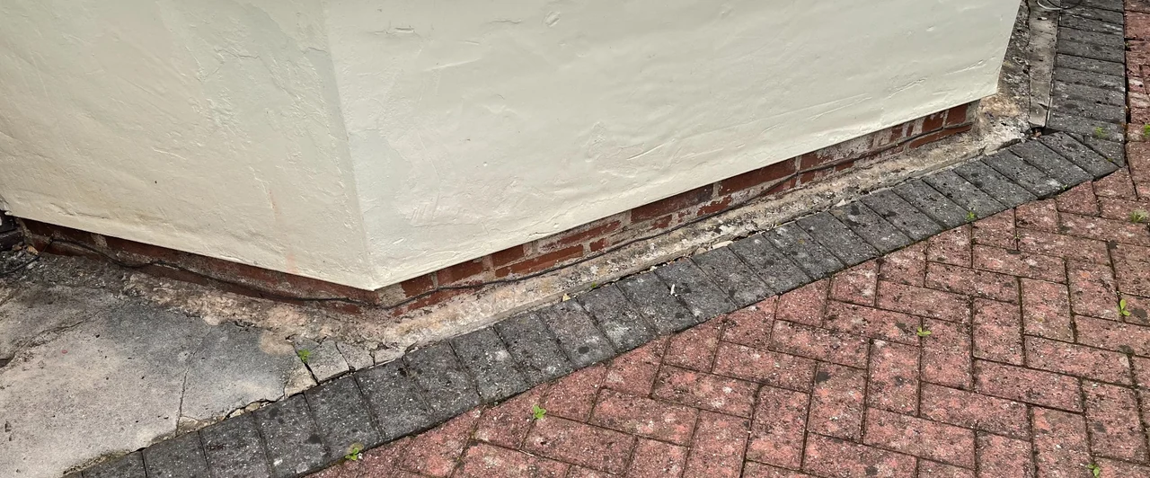Concrete visible along the base of a rendered bay wall beside block paving in Strines