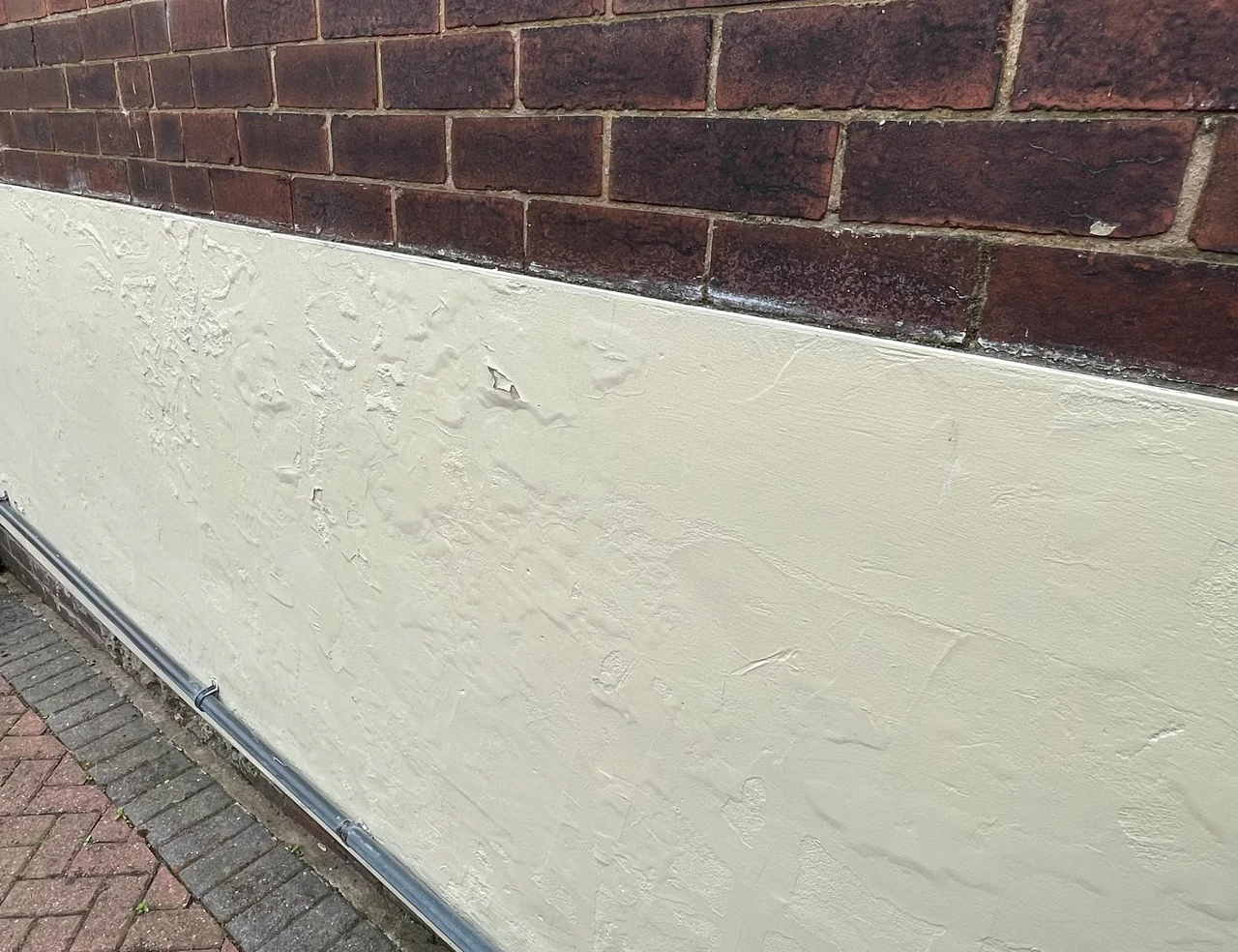 Deteriorating low-level rendered band beneath exposed brickwork in Strines